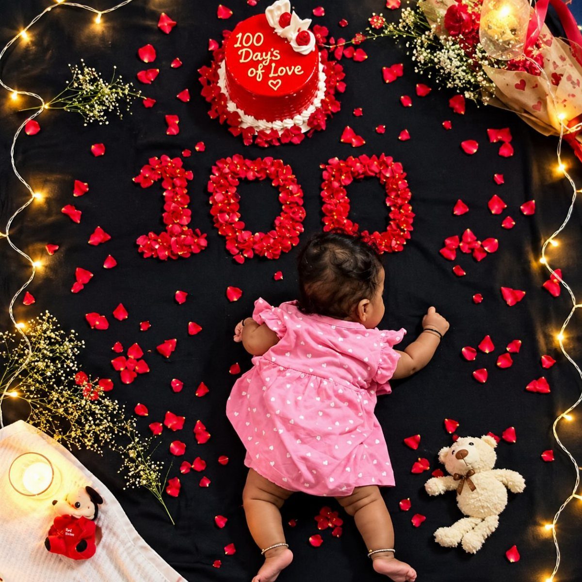 Baby in pink dress lying next to red rose petals forming number 100