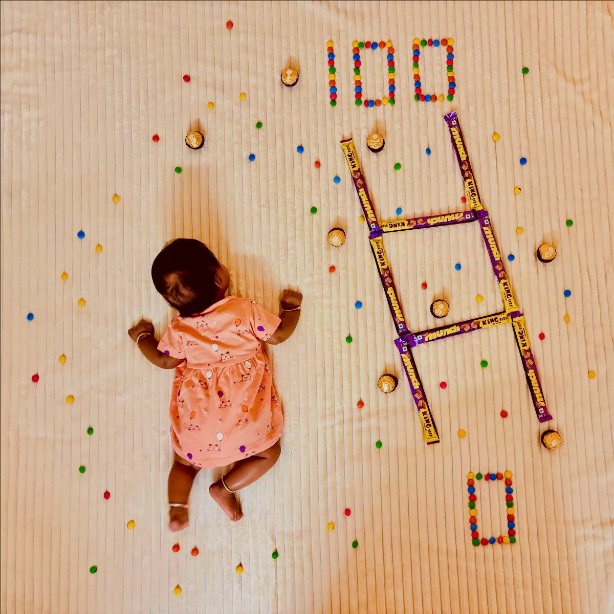Baby in pink dress lying next to 100 spelled with colorful candies