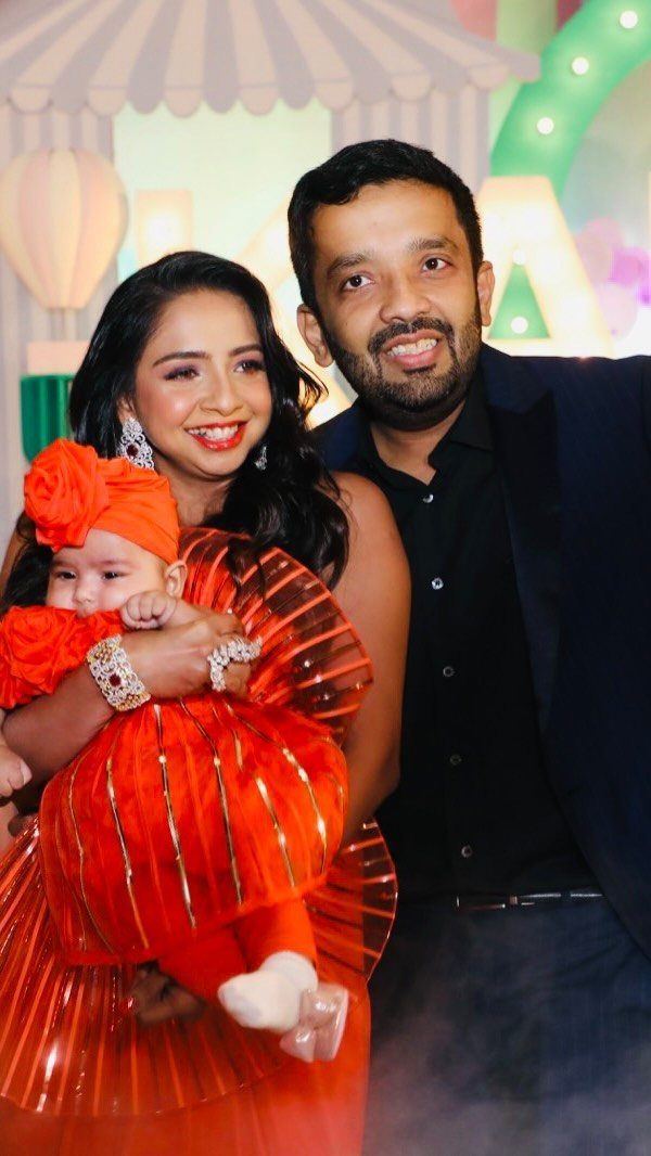 Baby in a bright orange ruffled dress and turban held by parents.