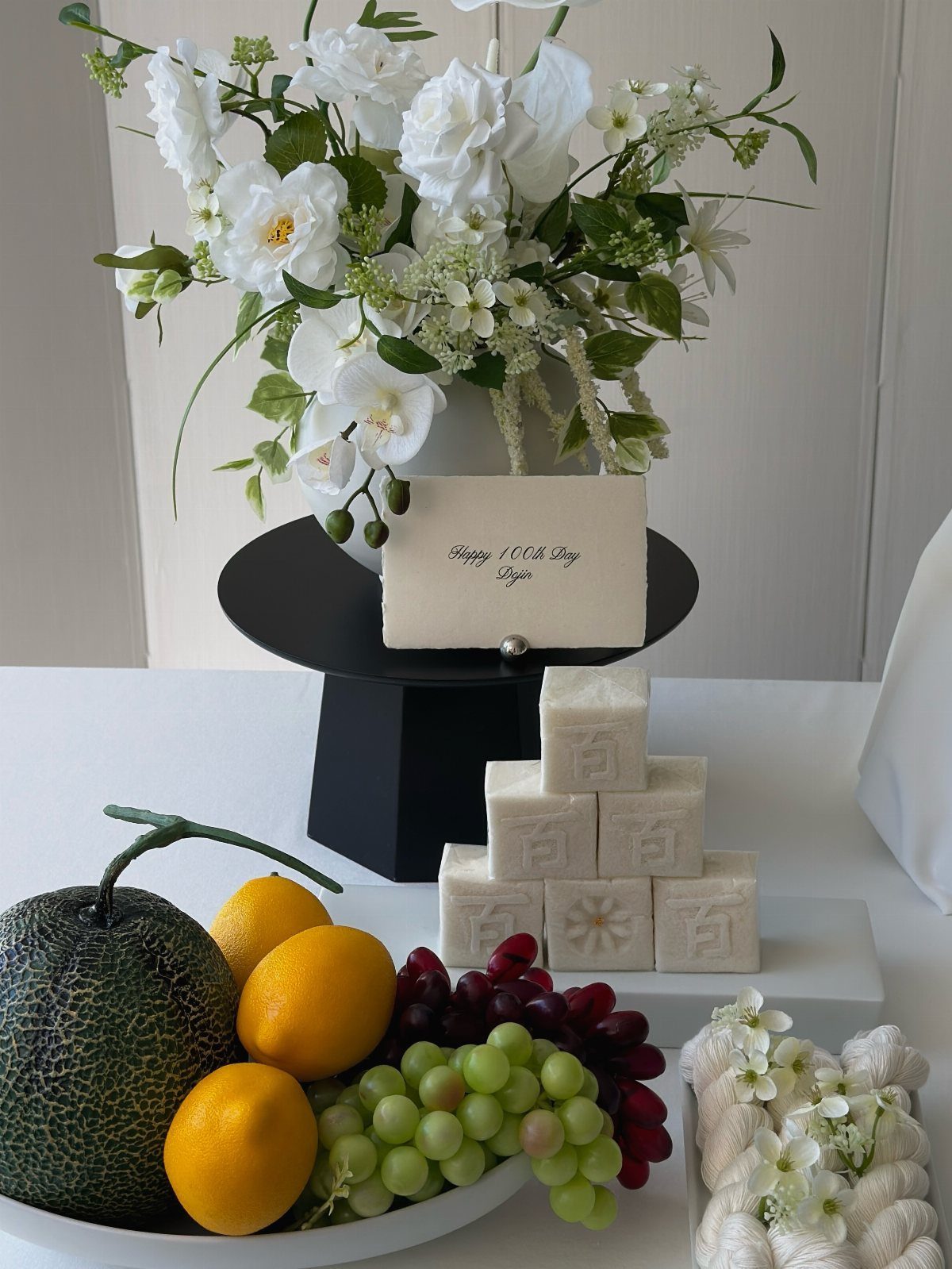 White flower bouquet, small cake, and fruit bowl on a white table.