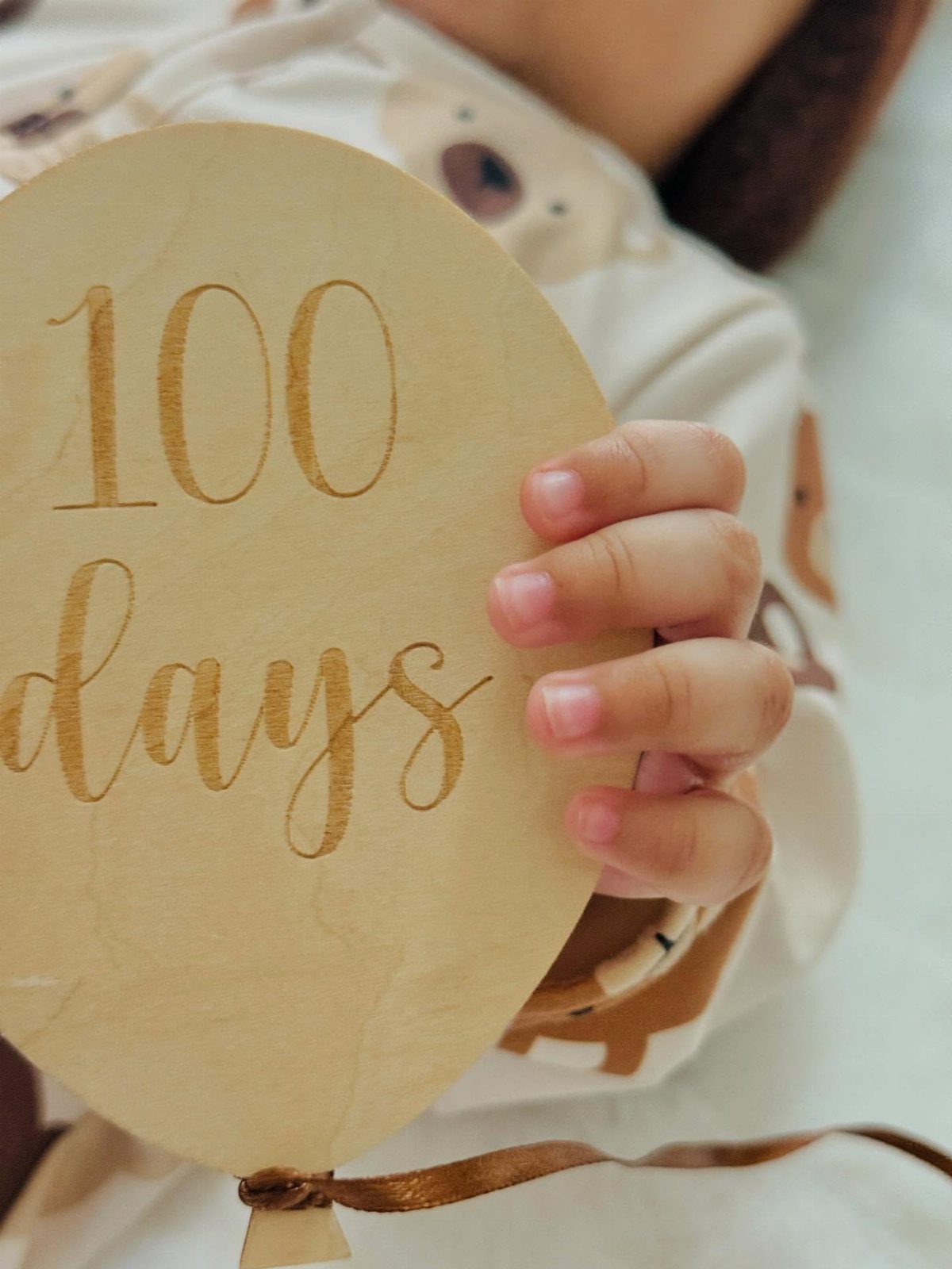 Baby's hand holding a wooden balloon plaque engraved with 100 days text