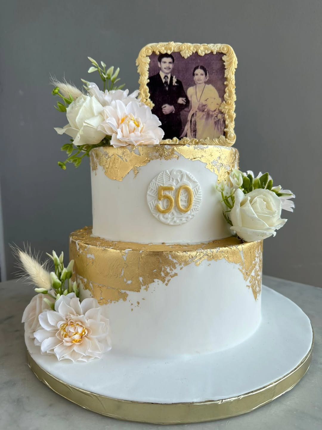 Two-tier white cake with gold leaf, white flowers, and a vintage couple's photo.