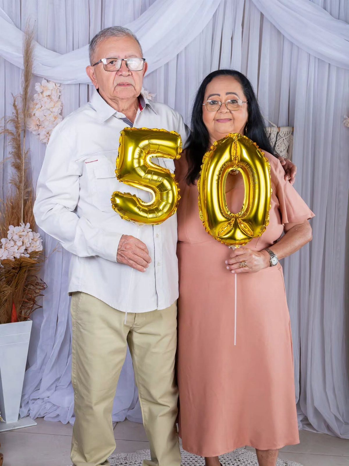 Older couple holding gold number 50 balloons in front of white curtains.