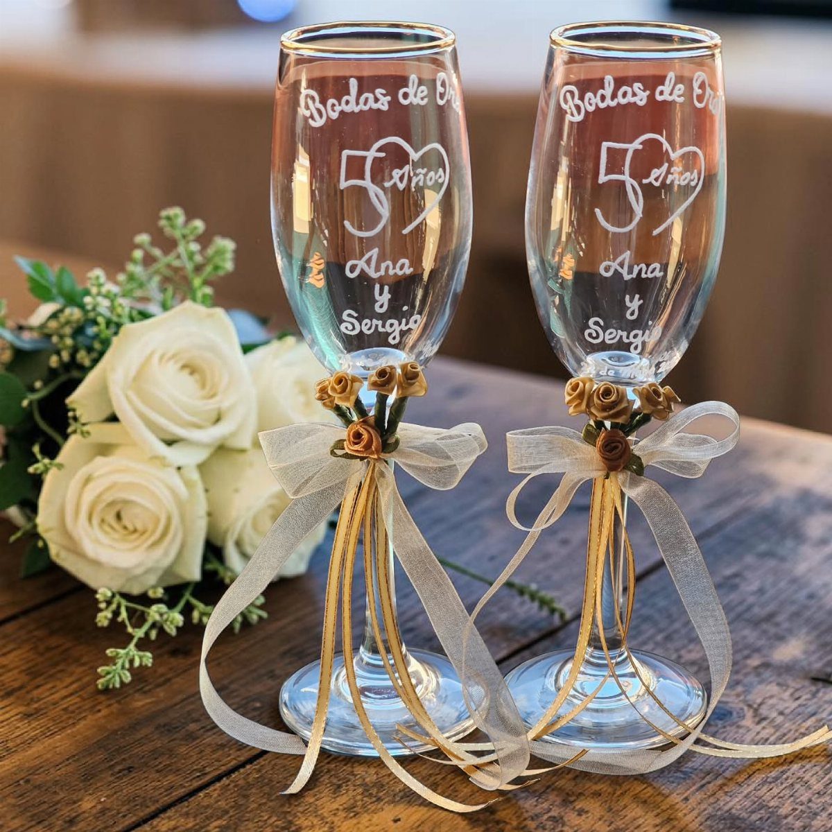 Two etched champagne flutes decorated with gold ribbons and roses beside white flowers.