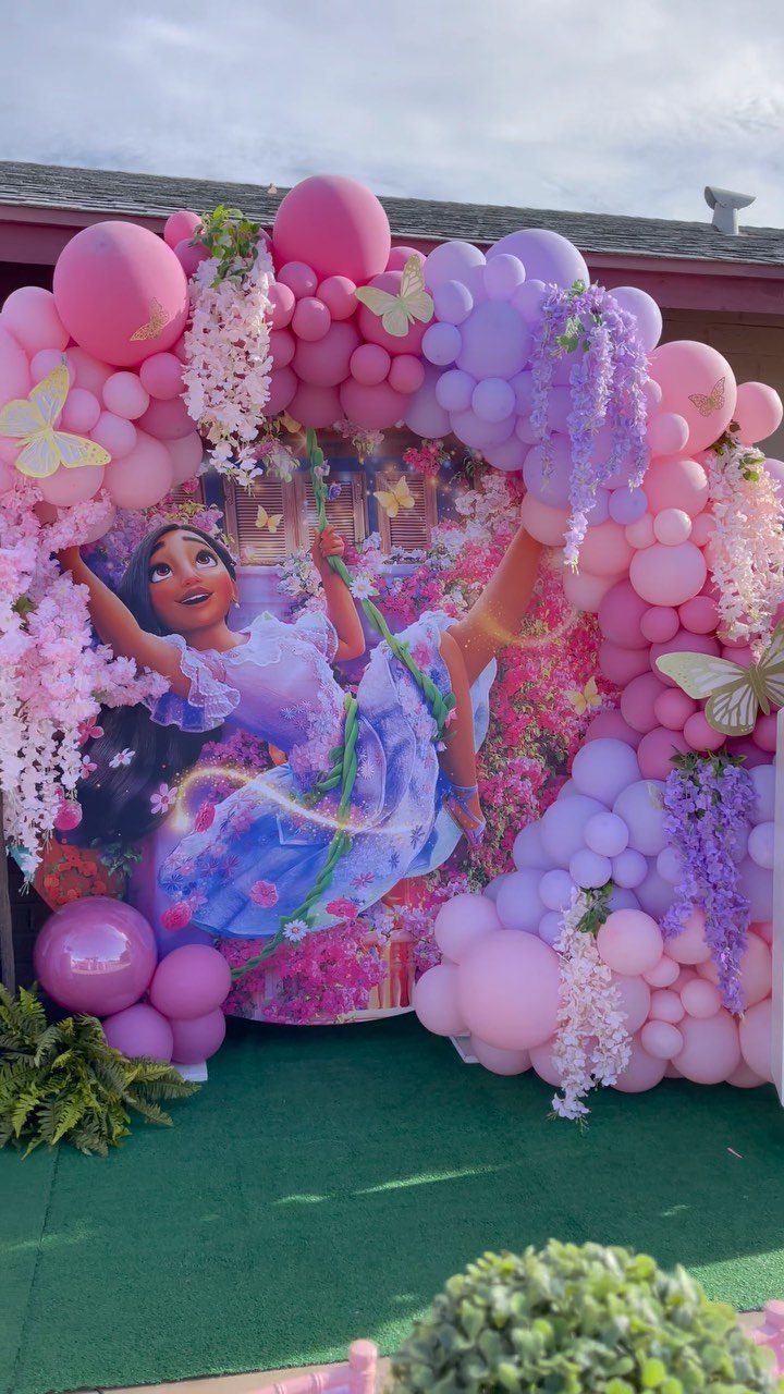 Encanto party backdrop with Isabela cutout, pink and purple balloons, and floral decorations.