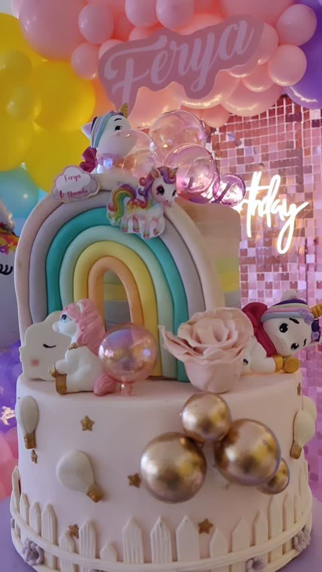 Pink unicorn cake with a rainbow, figurines, and balloons in the background.