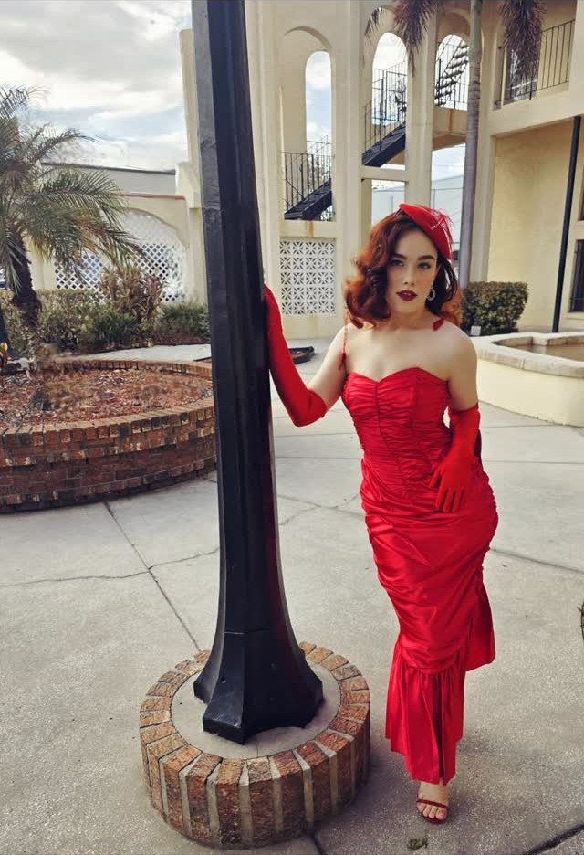 Woman in a red satin gown and gloves posing by a black lamp post.