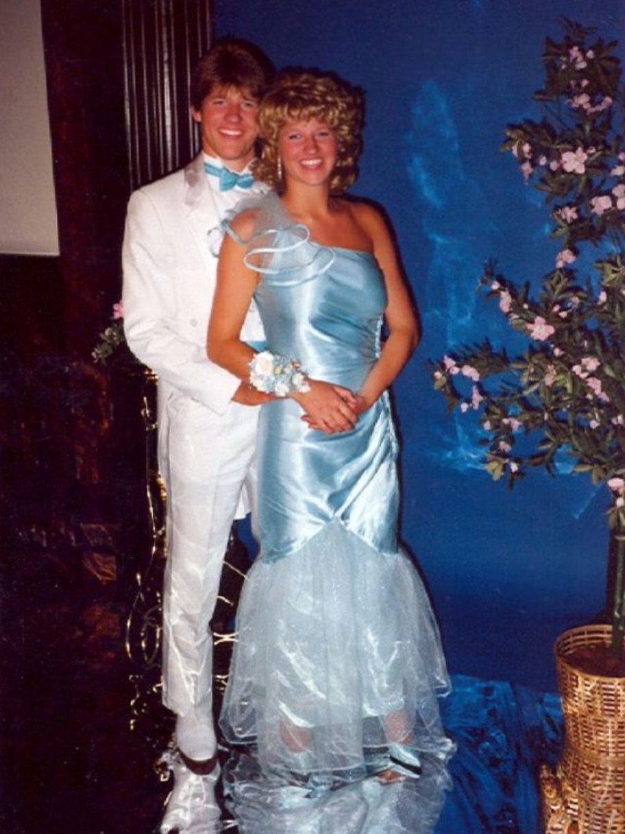 Young couple in 80s prom attire, man in white tuxedo and woman in blue gown.