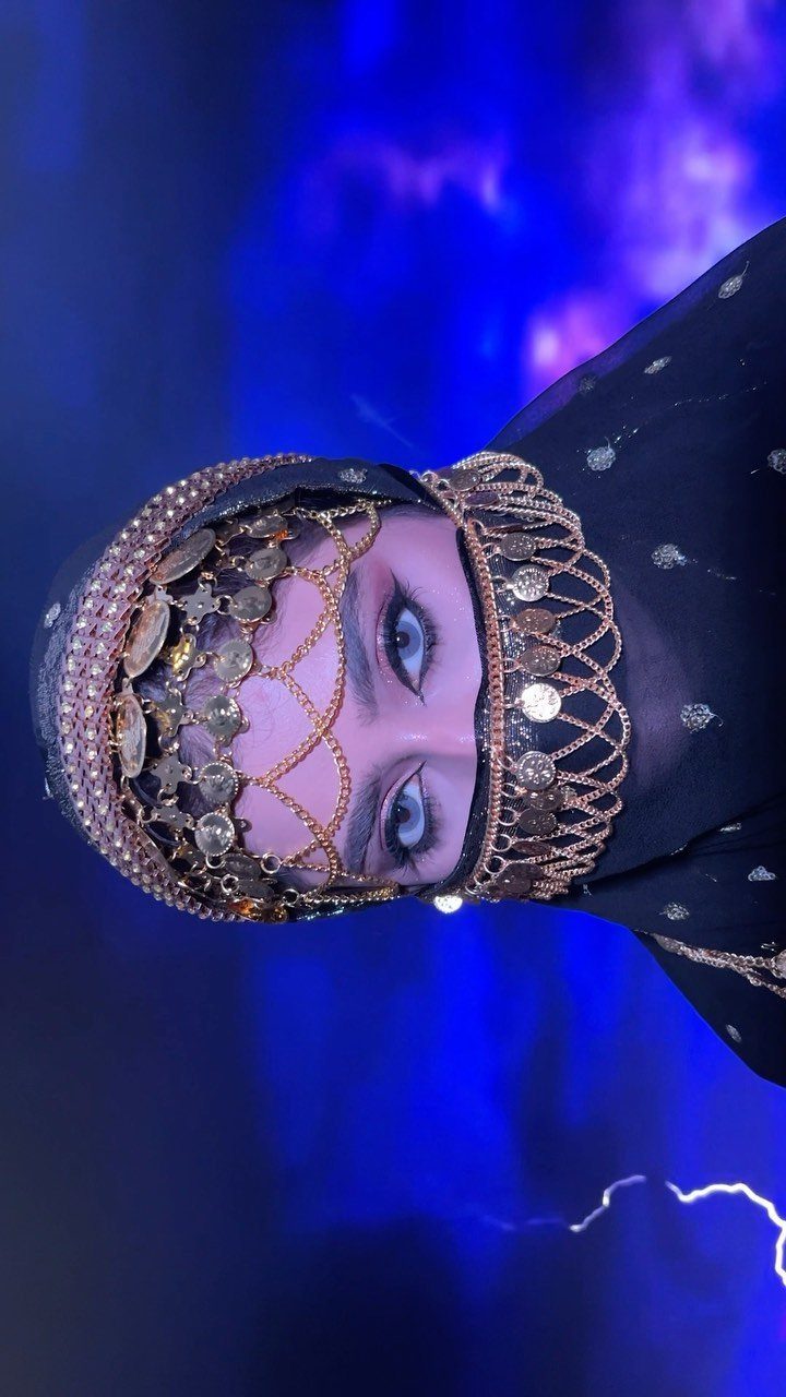 Person wearing an ornate gold coin headpiece and black veil against blue background