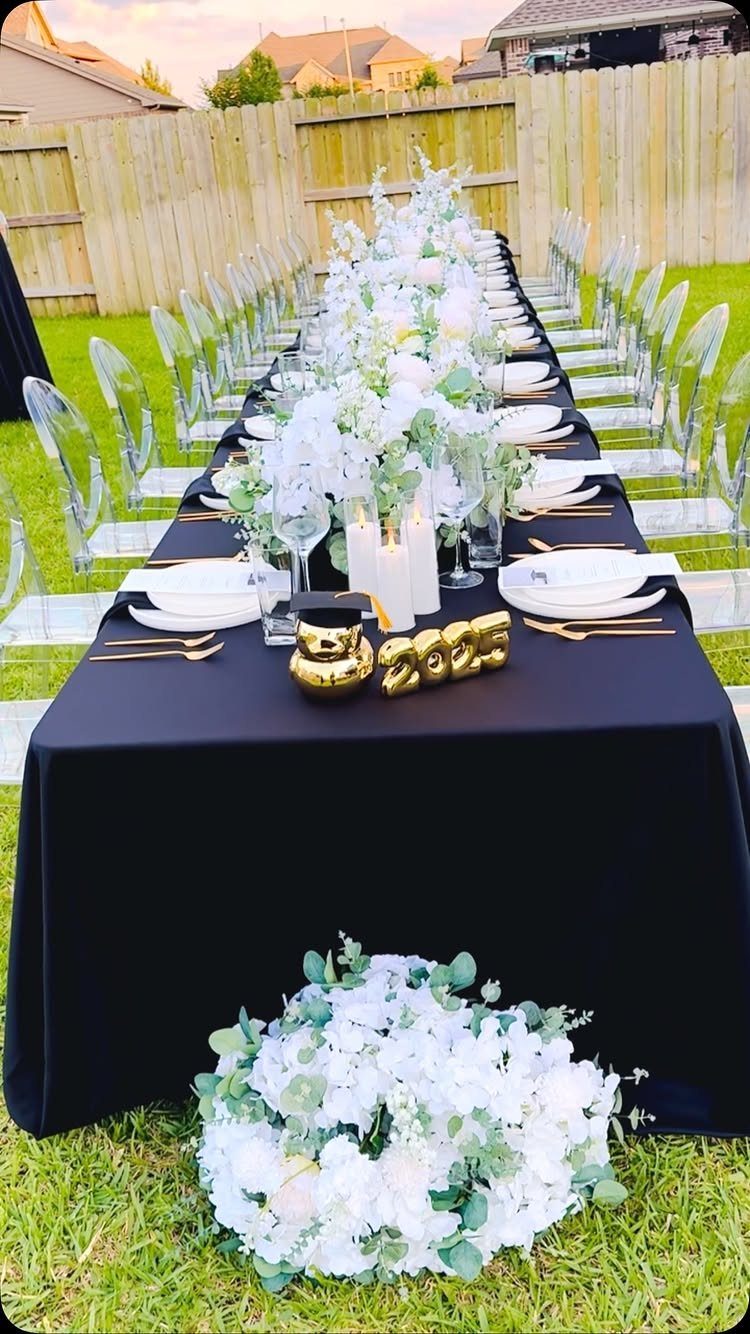 Long outdoor dining table with black cloth, white flowers, and gold graduation decorations.