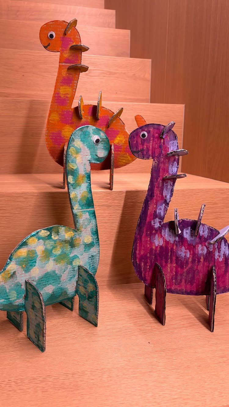 Three colorful painted cardboard dinosaur figures standing on light-colored wooden stairs.