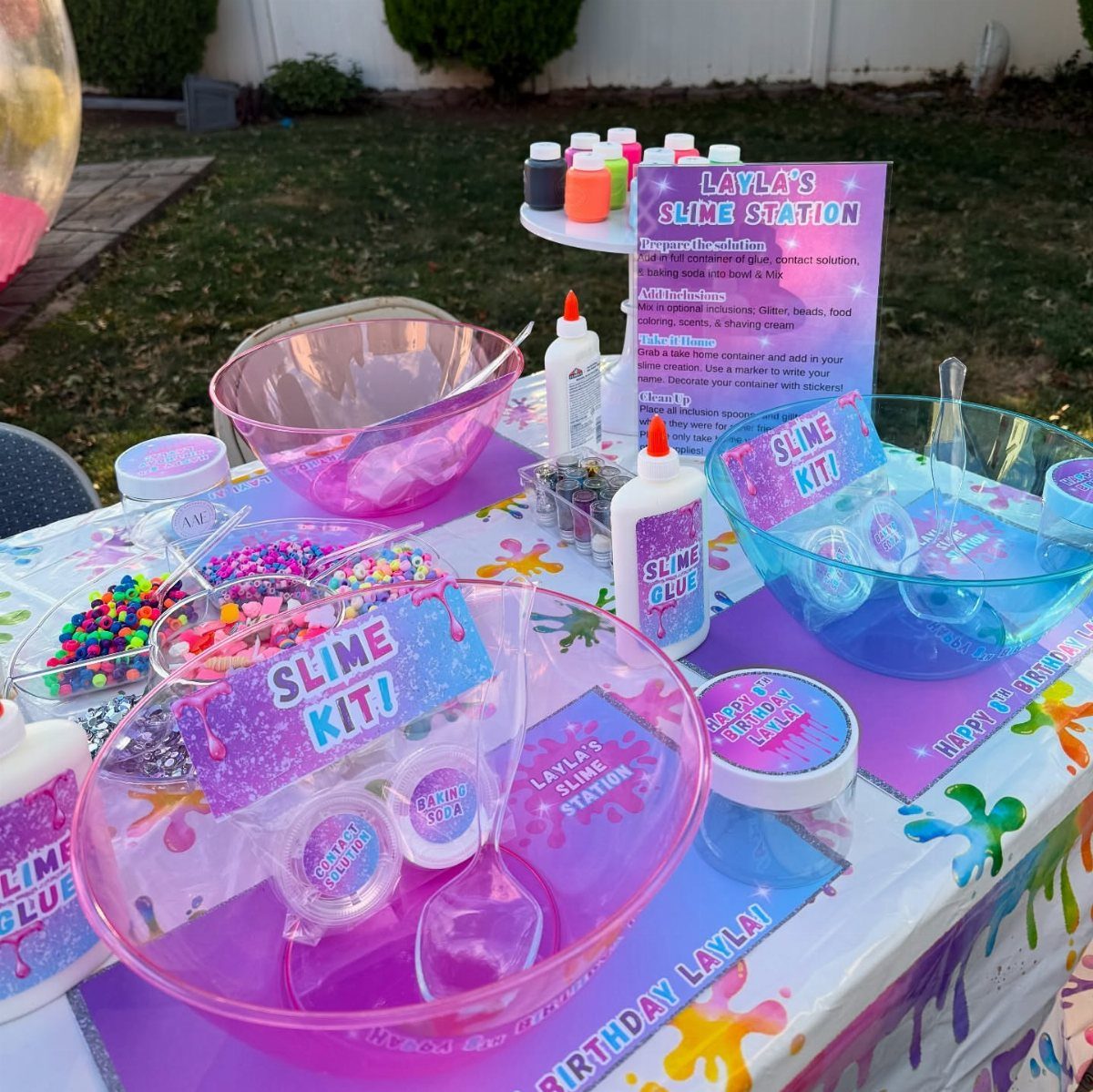 Slime making station with colorful bowls, glue, glitter, and instructional signs outdoors.