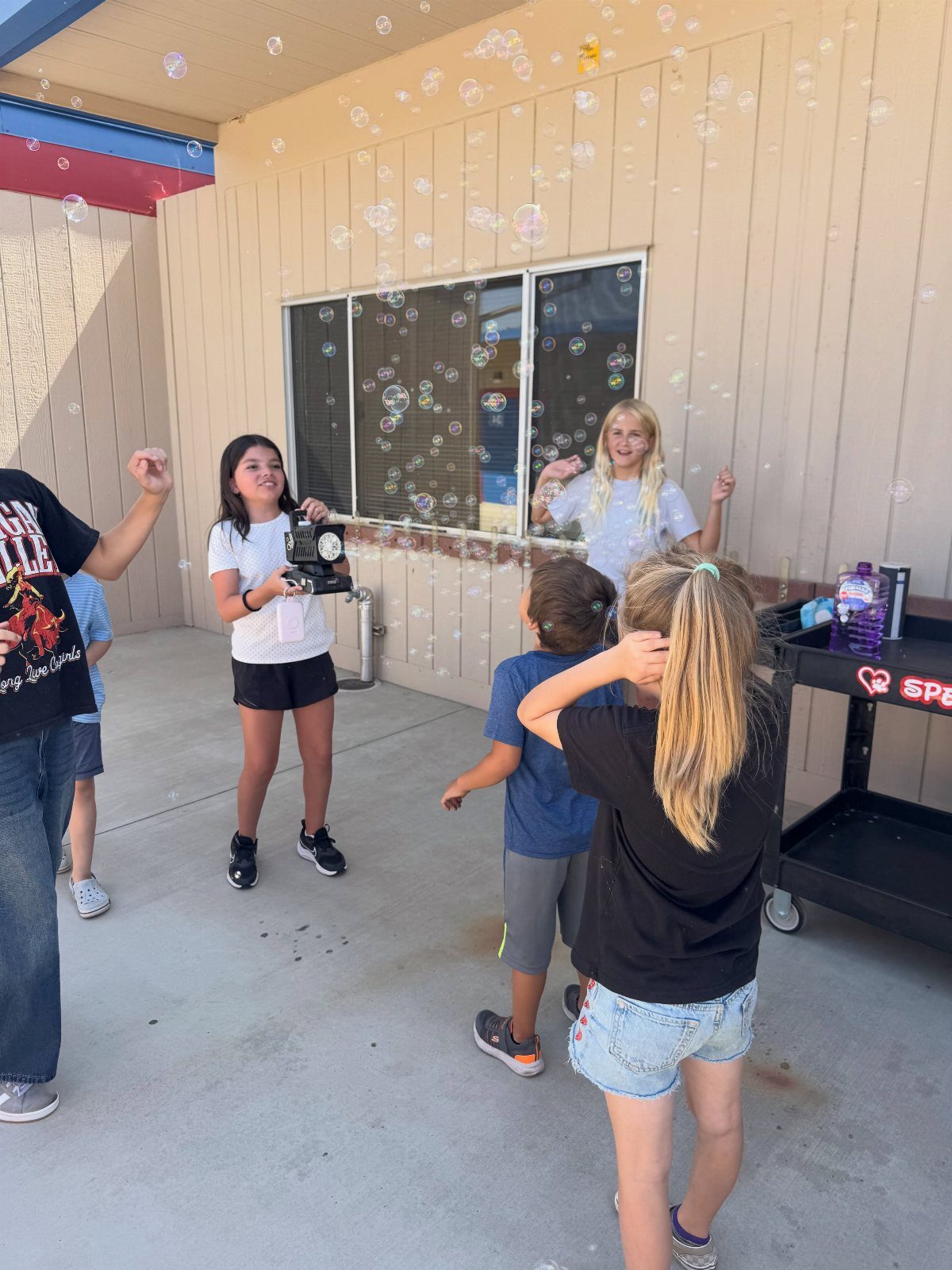 Children playing with a bubble machine outdoors in front of a school building.