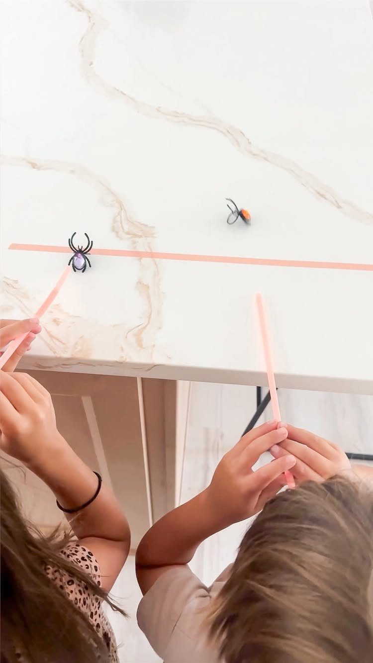 Children using pink straws to move plastic spiders across a marble table.
