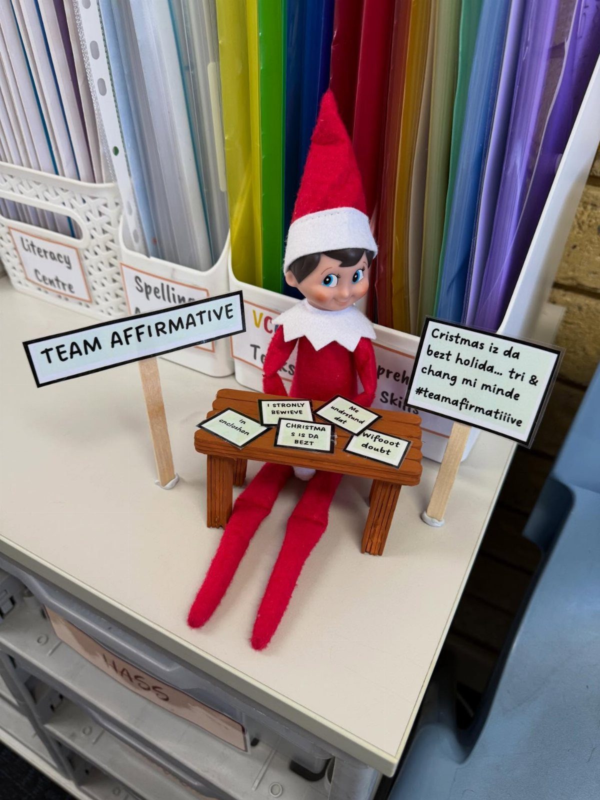 Elf on the Shelf doll sitting at a tiny desk in a classroom