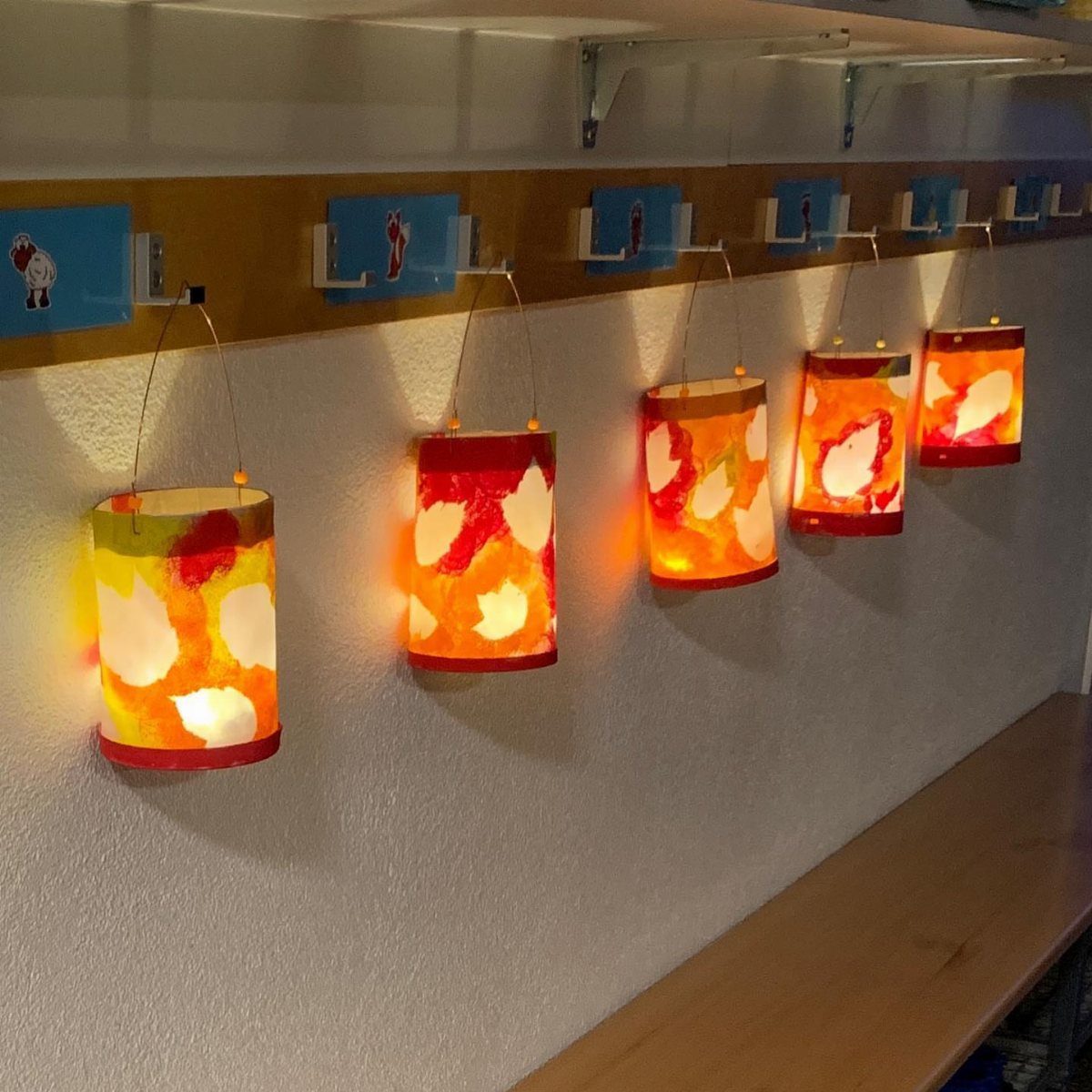 Colorful handmade paper lanterns glowing and hanging from a wooden ledge against a wall.