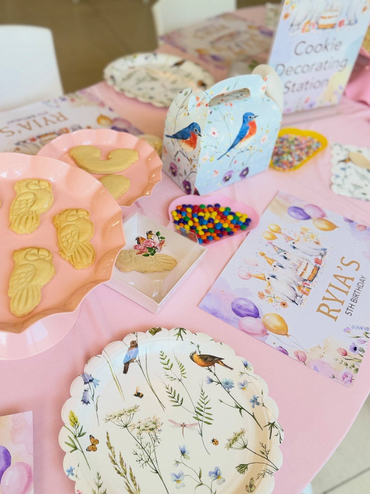 Cookie decorating station with plain cookies, colorful sprinkles, and bird-themed decorations on pink tablecloth.