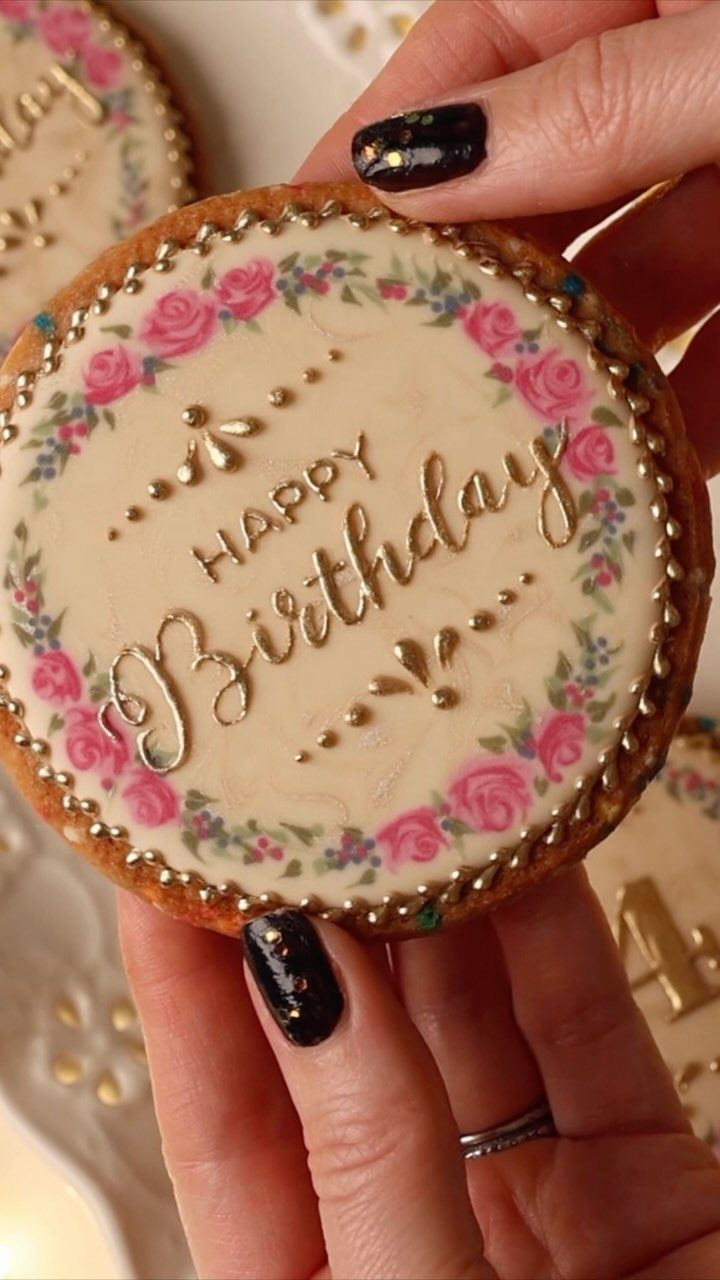 Decorated birthday cookie with pink roses and gold cursive lettering held by hands.