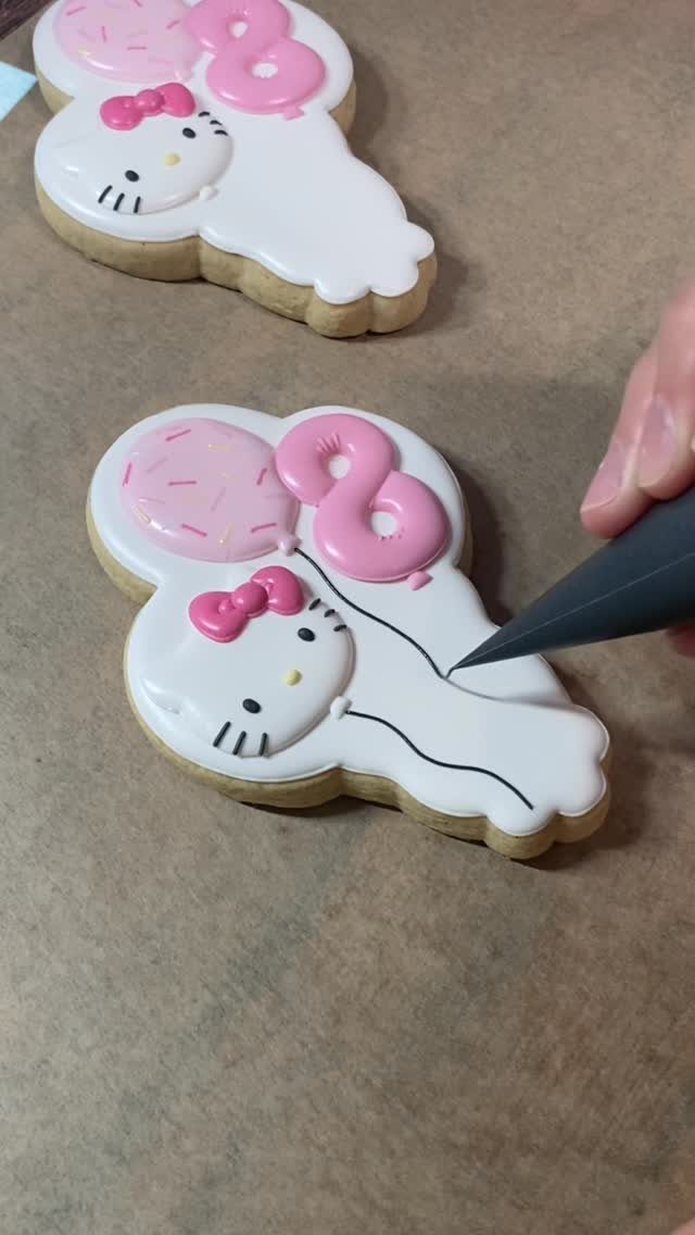 Hand piping black icing onto a Hello Kitty shaped sugar cookie with pink balloons