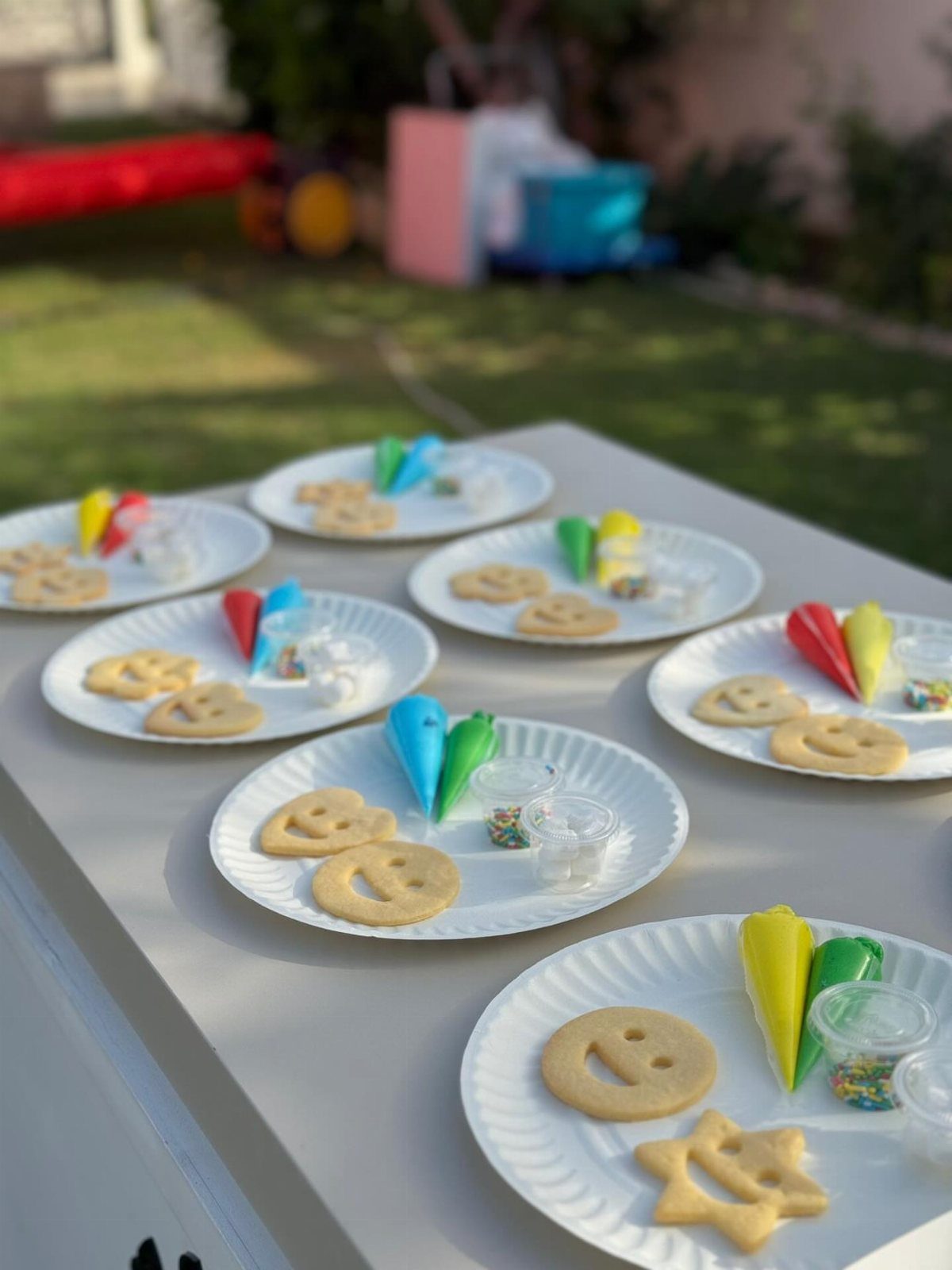 Plates of plain sugar cookies with colorful icing bags and sprinkles on a table.
