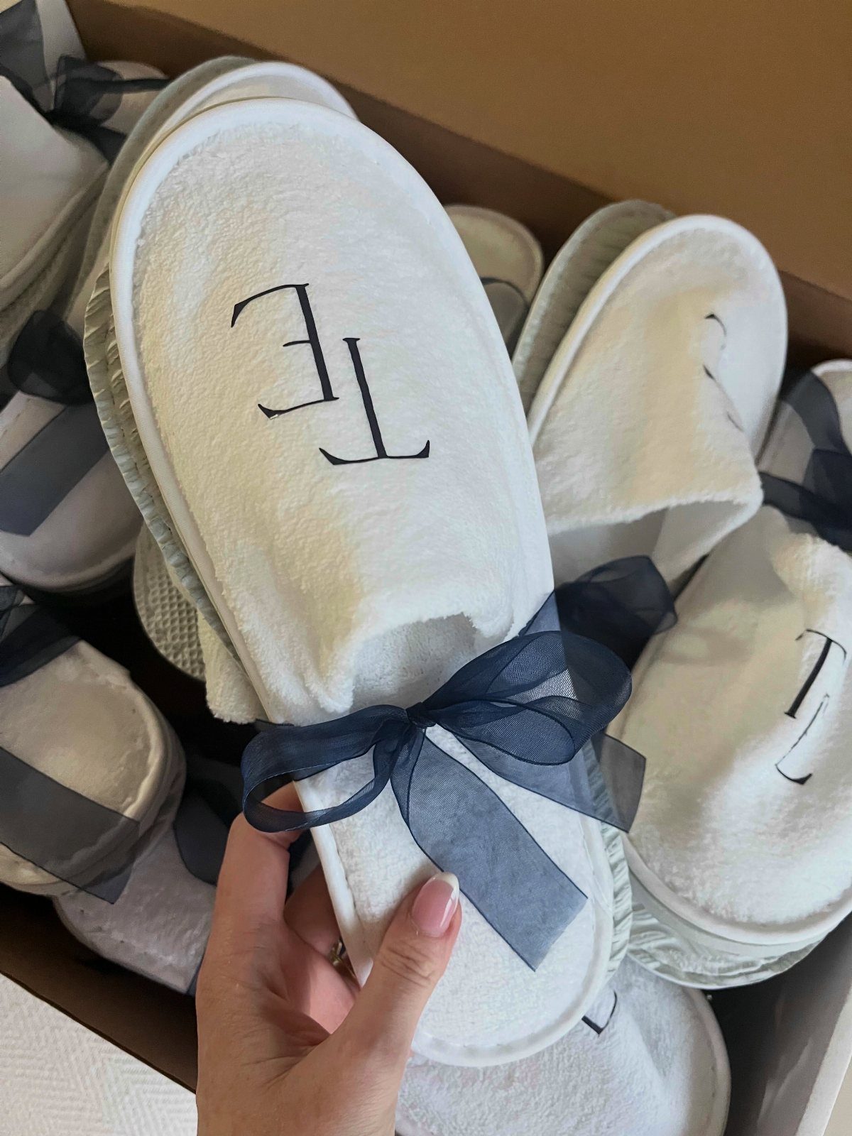 White plush slippers with embroidered initials and navy blue ribbons in a box