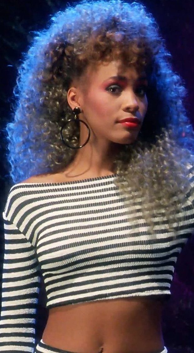 Woman with big curly hair and striped crop top in 80s style attire.