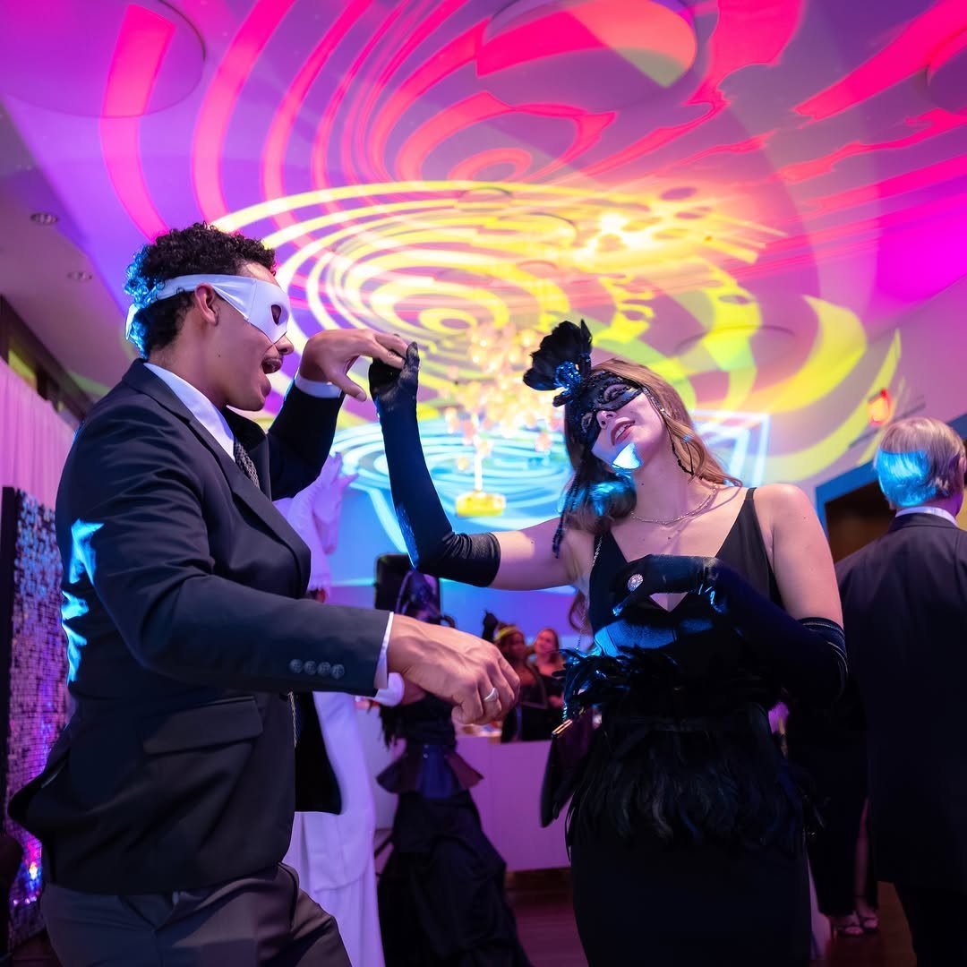 Couple in formal attire and masquerade masks dancing under colorful swirling ceiling lights.
