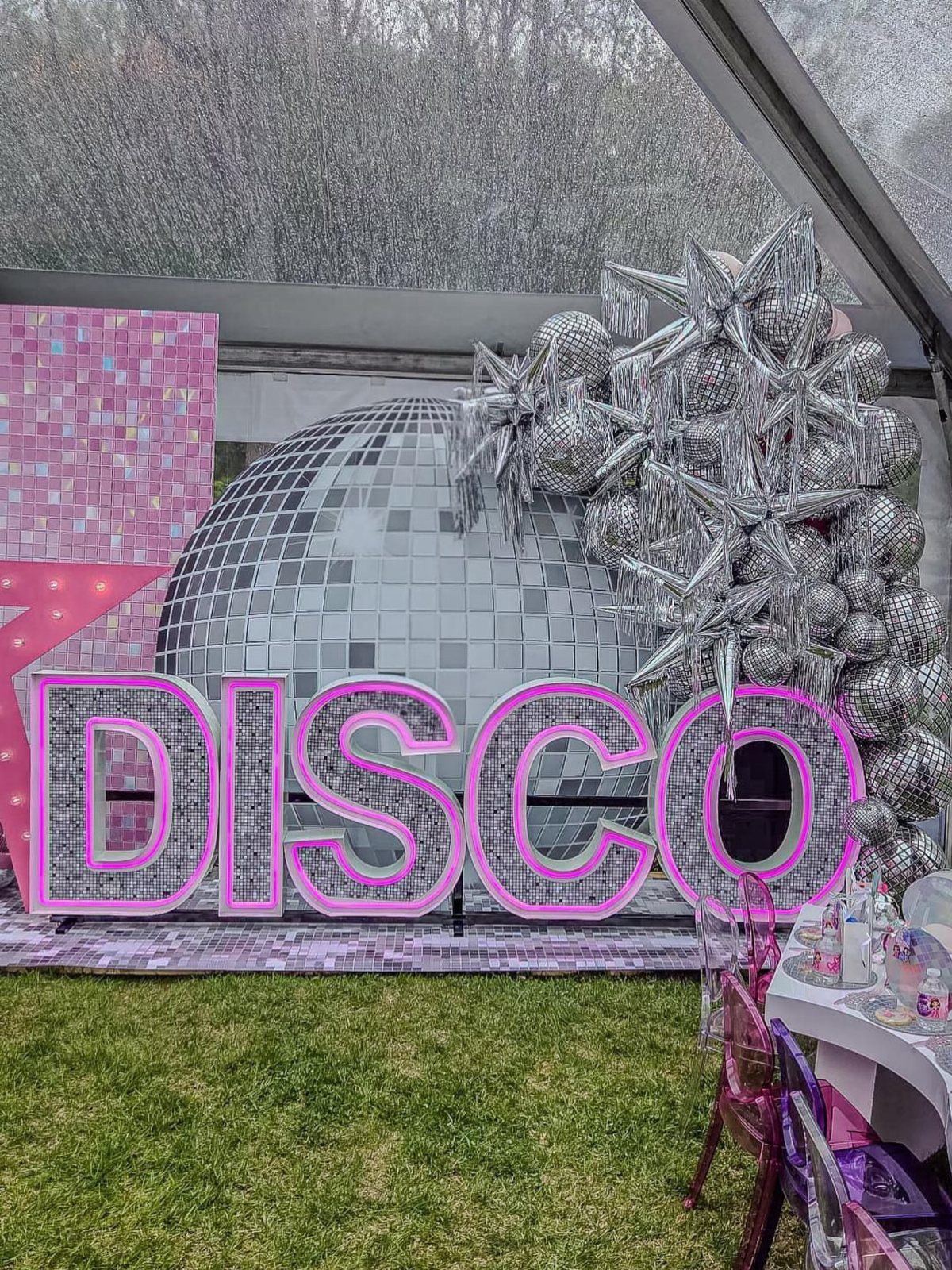 Large silver disco ball backdrop with glowing pink DISCO letters on green grass.