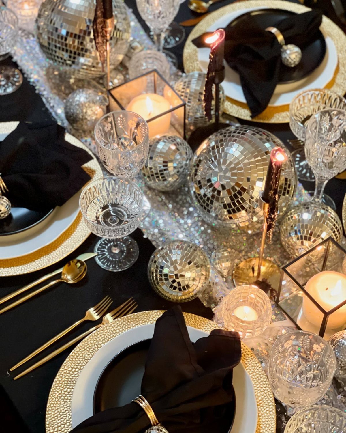 Dinner table setting with black plates, gold cutlery, and silver disco ball centerpieces.