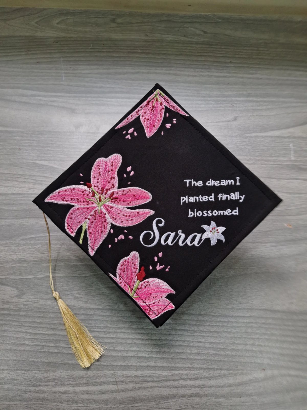 Decorated black graduation cap with pink lilies and inspirational text on grey wood.