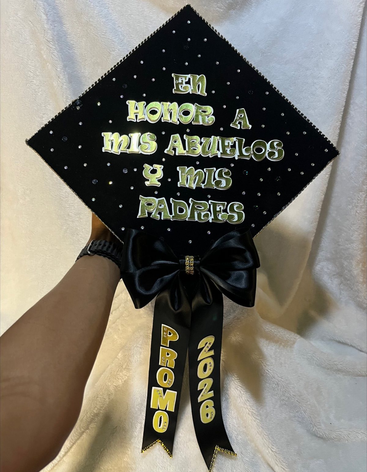 Decorated black graduation cap with gold Spanish text and a large satin bow.