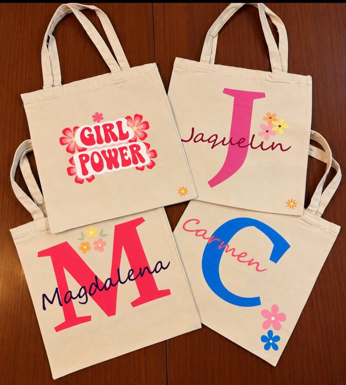 Four beige canvas tote bags with colorful monograms and names on wooden background