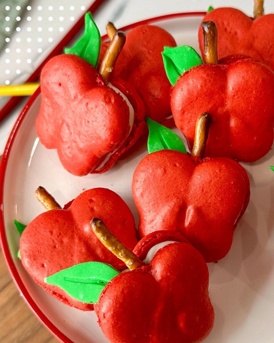 Apple-shaped red macarons with green leaves and pretzel stems on a white plate.