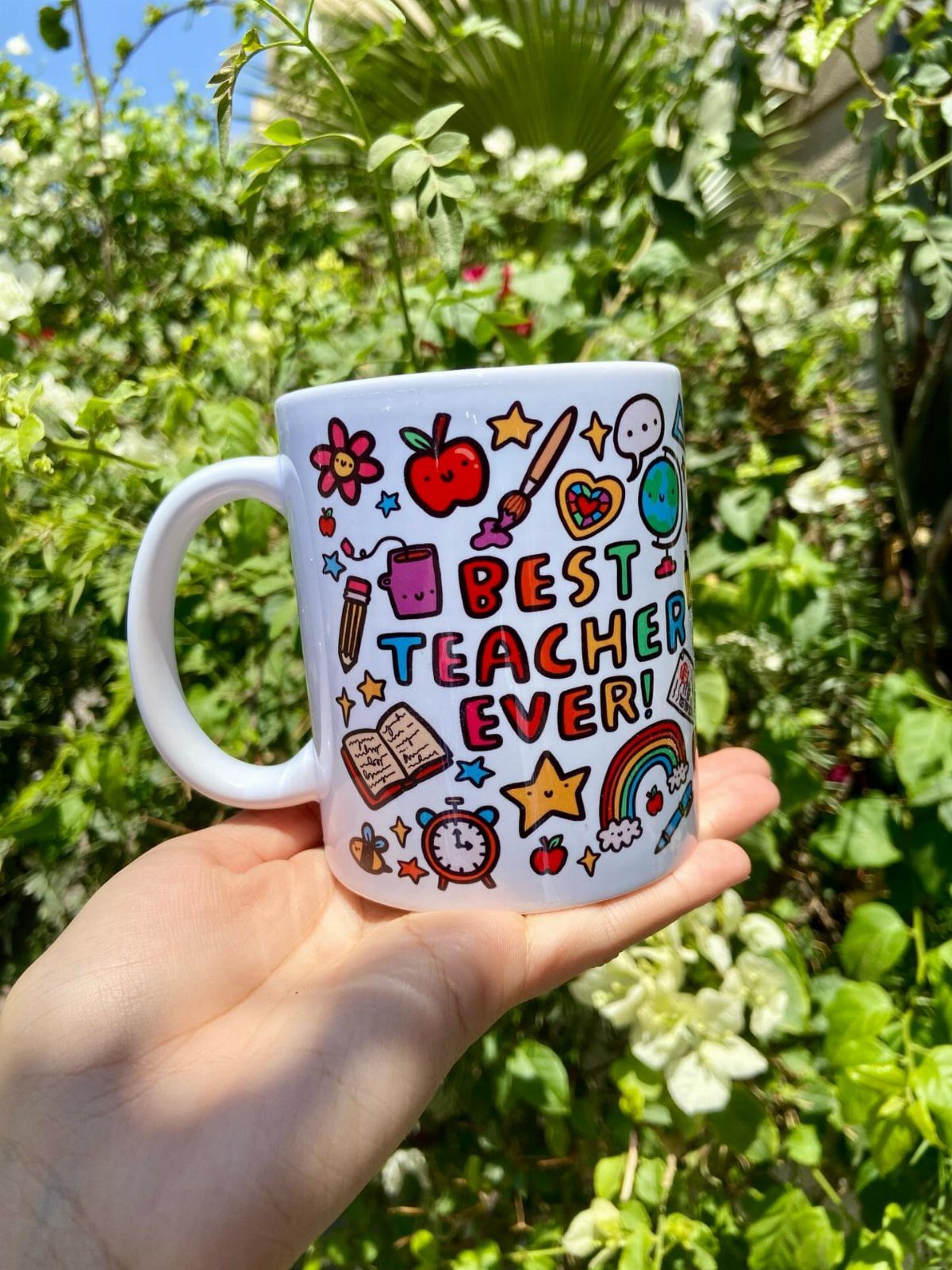 White ceramic mug with colorful teacher-themed drawings and text held against green foliage.