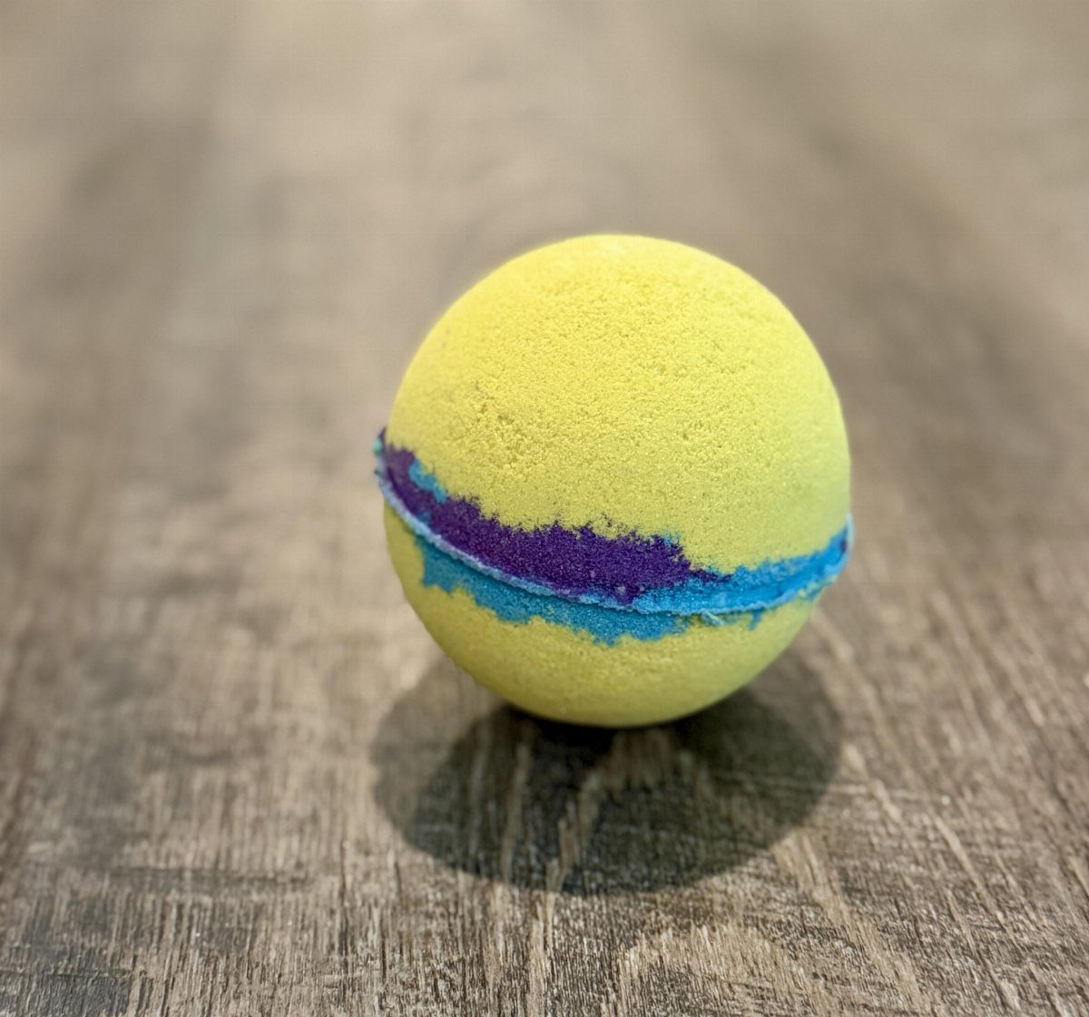Yellow bath bomb with blue and purple stripes on a grey wooden surface.