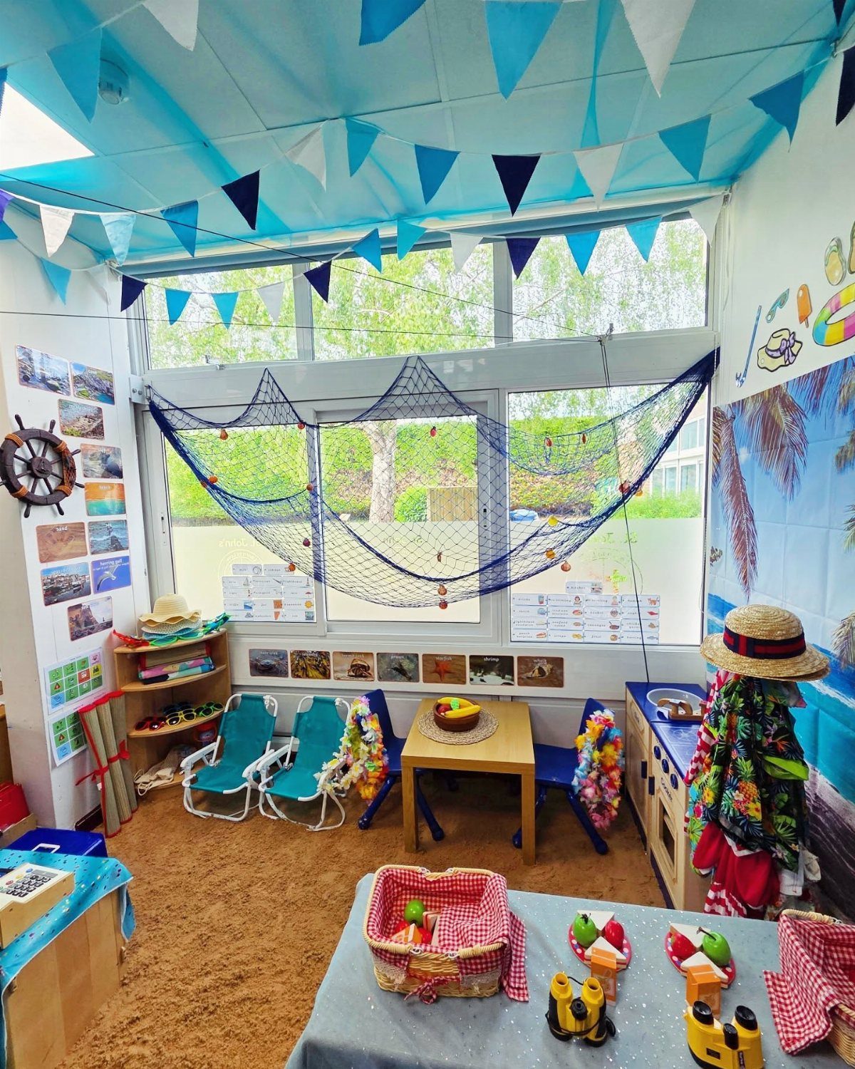 Beach themed classroom with blue bunting, fishing nets, lounge chairs, and sand flooring.