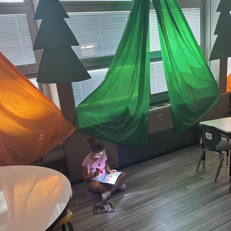 Young girl reading a book inside a camping themed classroom with tents and trees