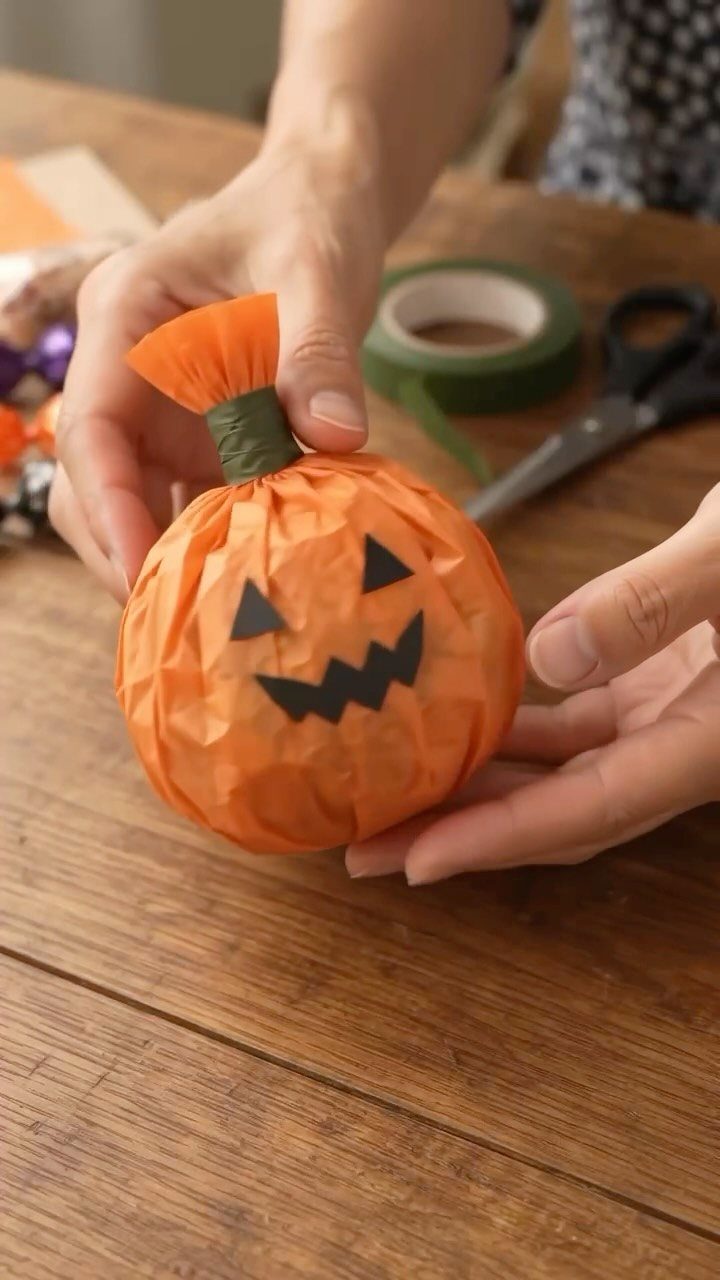 Hands holding a small orange tissue paper pumpkin with a black jack-o'-lantern face.