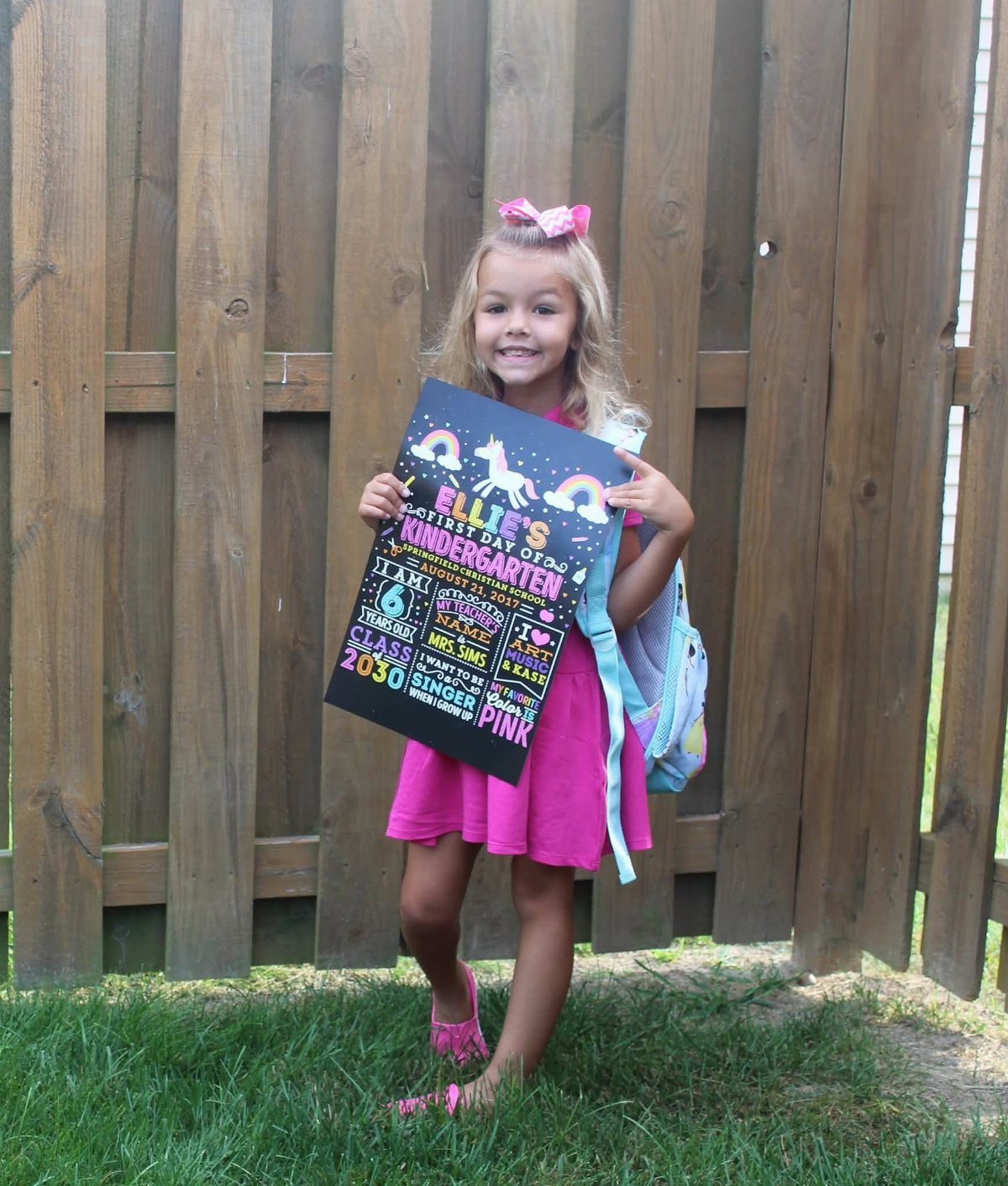Young girl in pink dress holding a first day of kindergarten chalkboard sign.