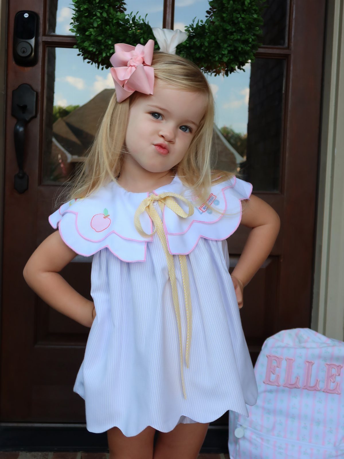 Young blonde girl in a white apple-themed dress posing before a brown door.
