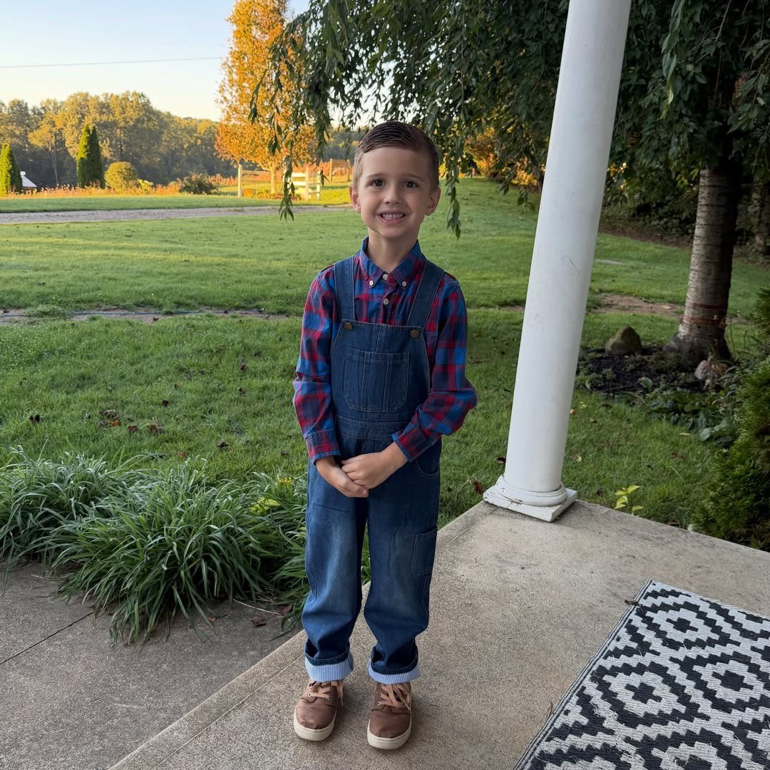 Young boy wearing blue denim overalls and a red plaid shirt on a porch