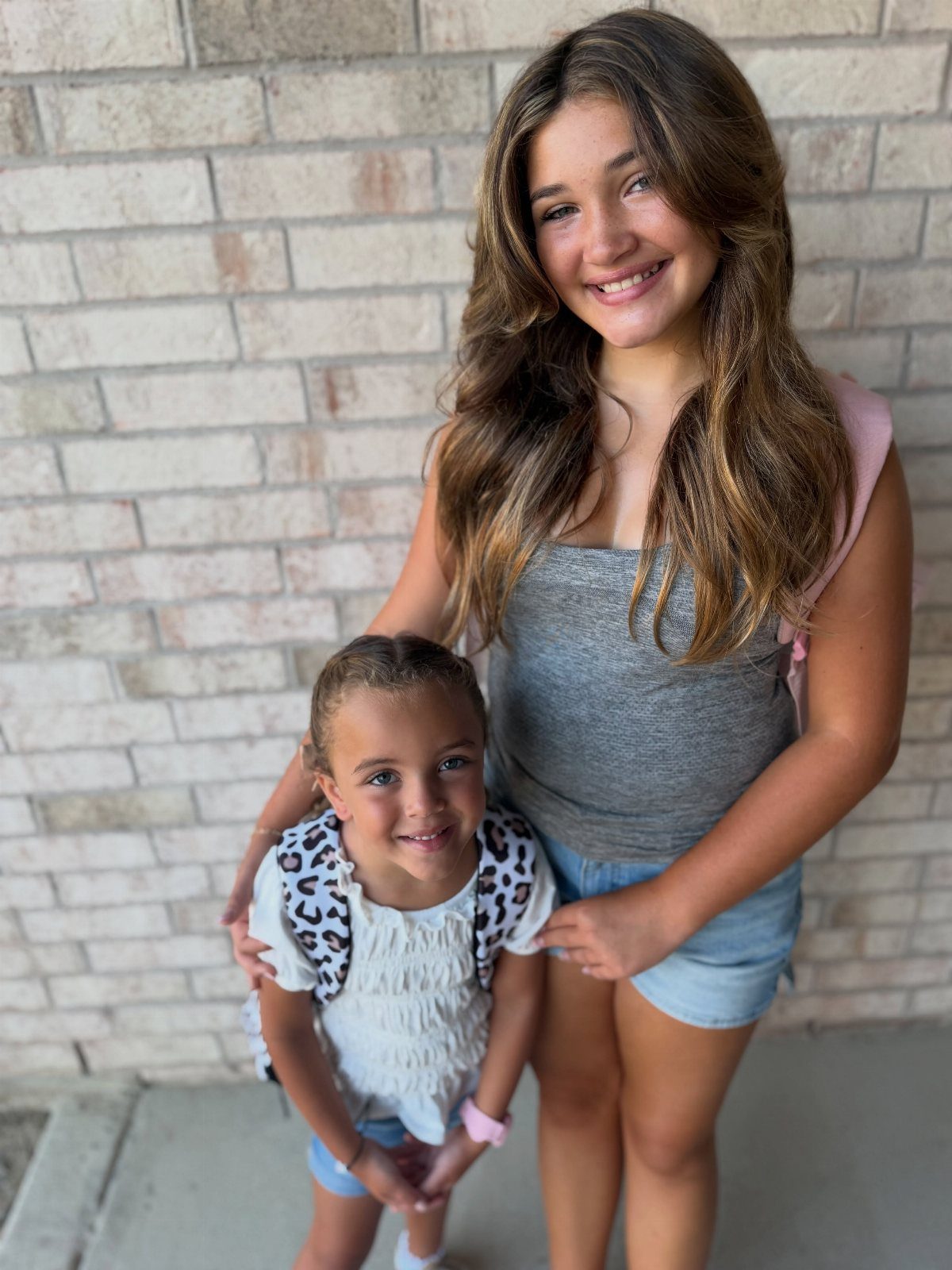 Teenage girl and young girl with leopard print backpack posing by brick wall