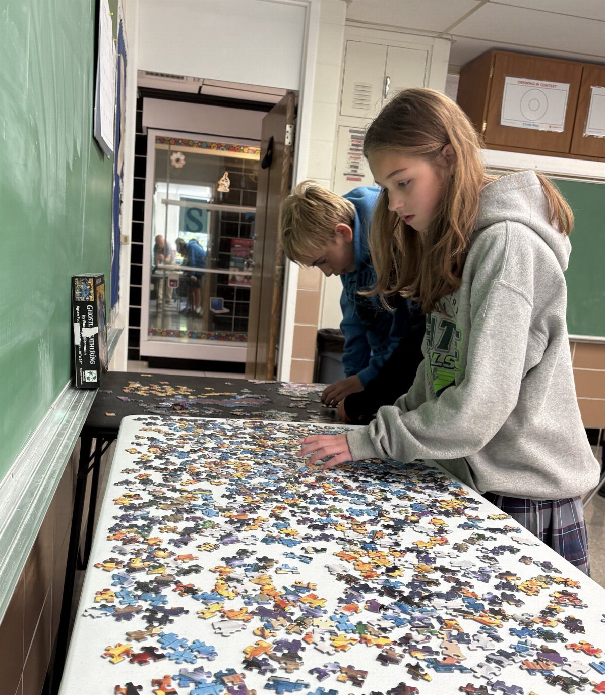 Two students assembling a large colorful jigsaw puzzle on a white classroom table.