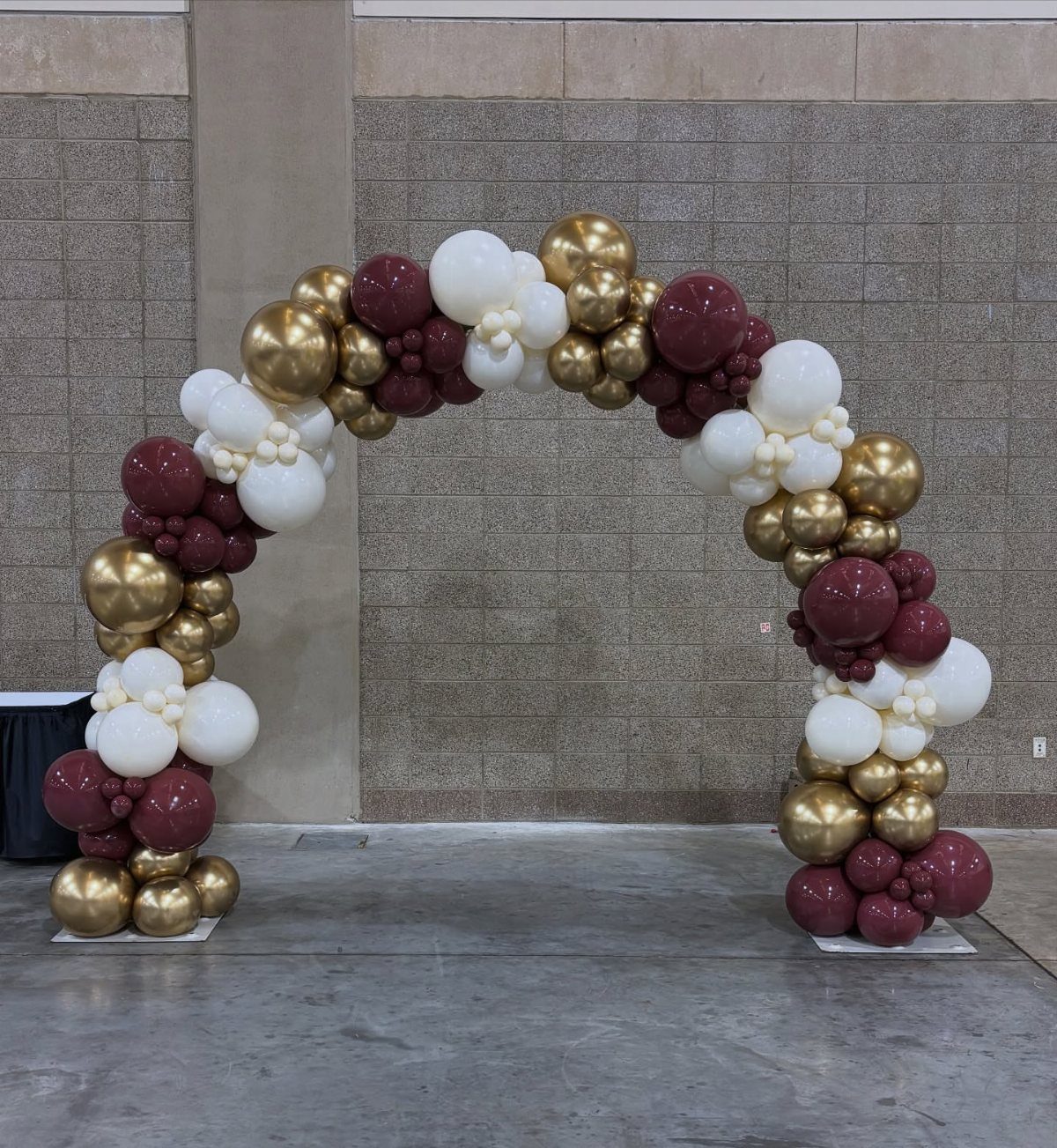Balloon arch in burgundy, gold, and white colors against a grey wall.