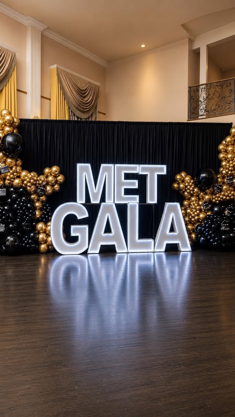 Illuminated MET GALA letters with gold and black balloon decor against black curtains.