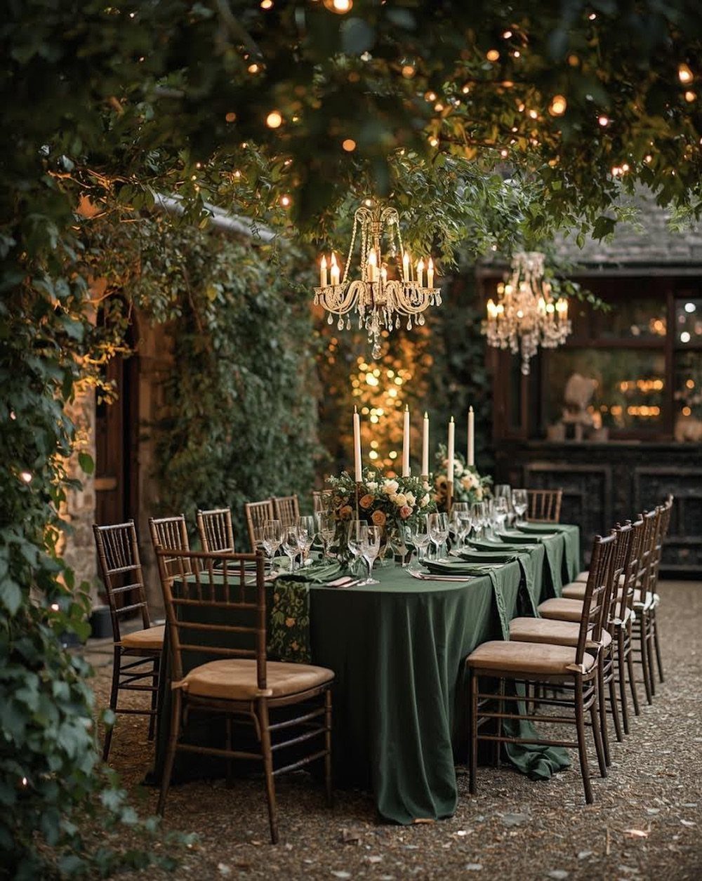 Long banquet table with green linens under a canopy of trees and chandeliers.