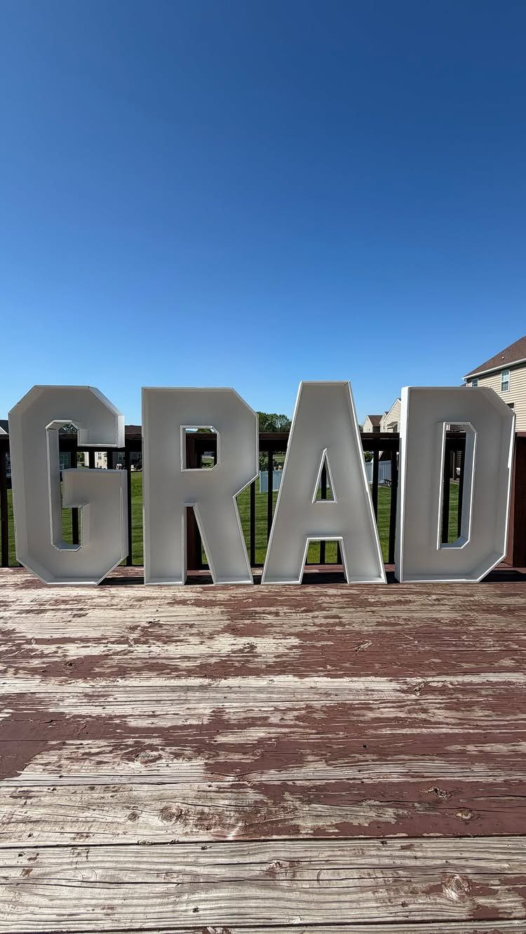 Large white GRAD block letters standing on a weathered wooden outdoor deck.