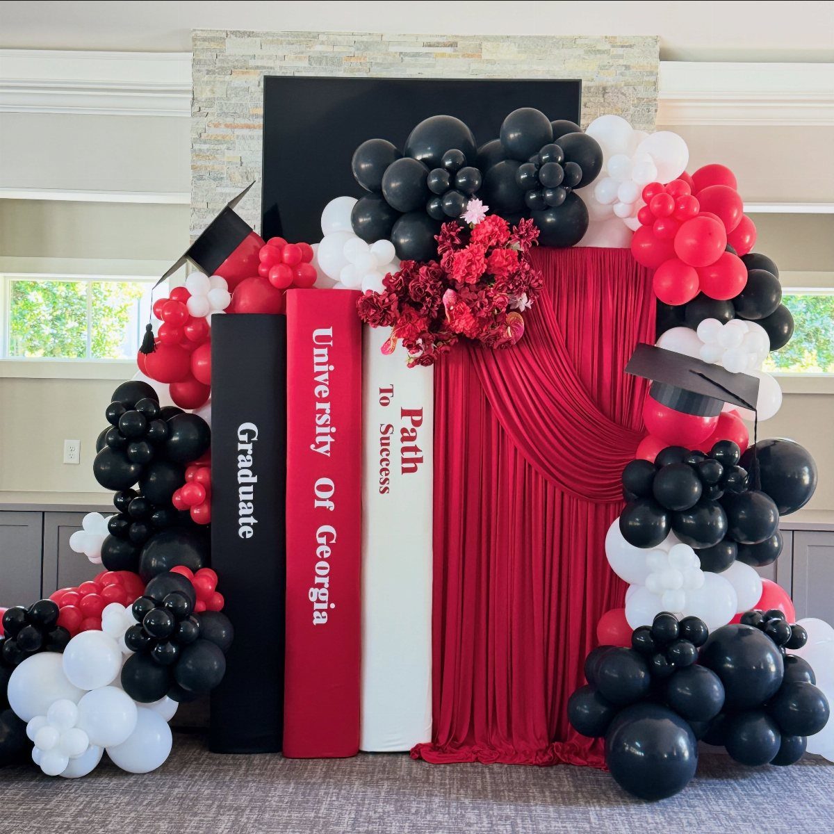 Graduation backdrop with oversized books, red fabric draping, and red, black, and white balloons.