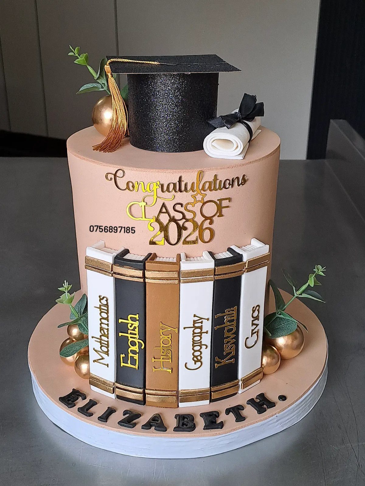 Two-tier peach graduation cake with a black cap, diploma, and edible school books.