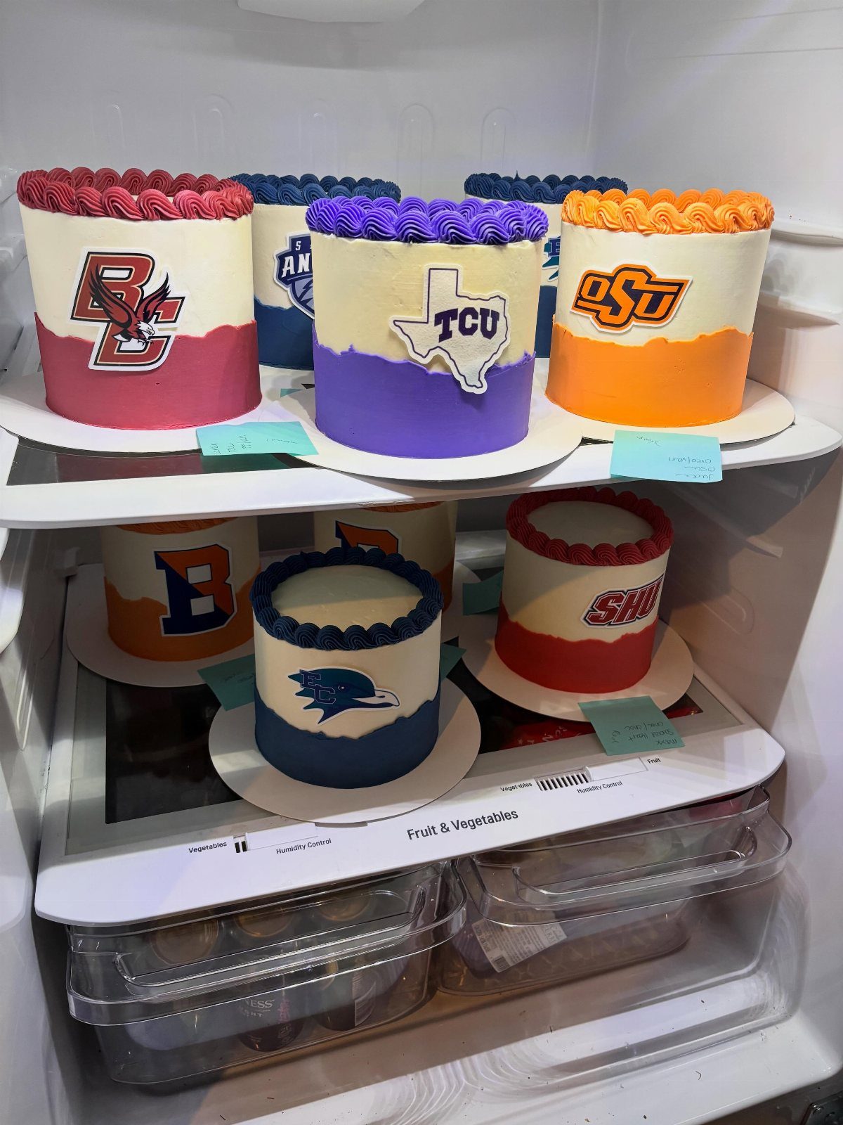 College logo cakes with colorful frosting displayed on refrigerator shelves.